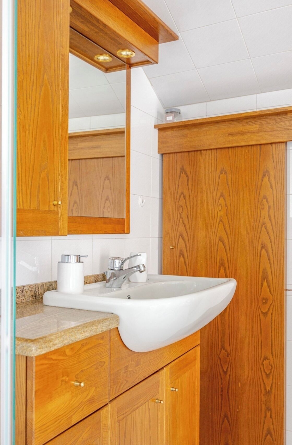 A wooden bathroom vanity with a white sink, a mirror, and a towel dispenser.