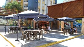 Un área de estar al aire libre con mesas y sillas bajo sombrillas, una pérgola de madera y vistas a un edificio alto al fondo.