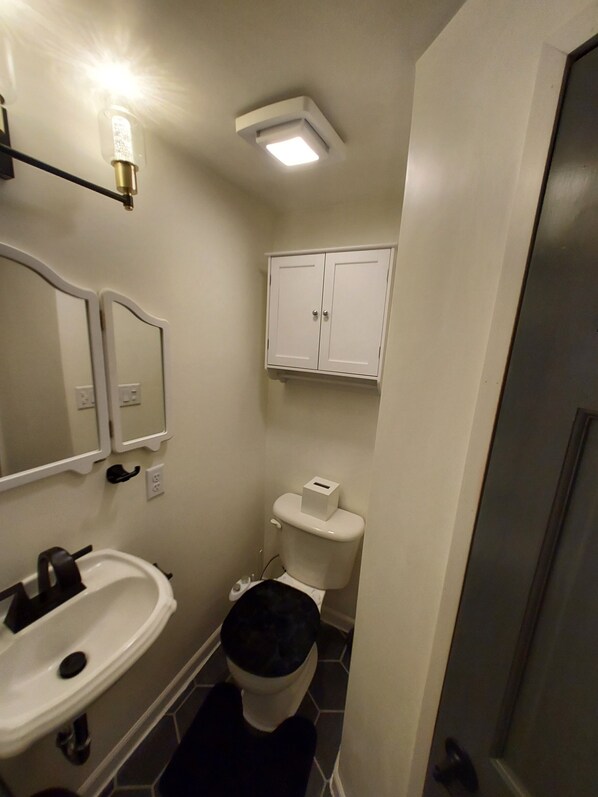 A small bathroom with a white sink, a toilet, and a mirror. There is a cabinet above the toilet and a light fixture on the ceiling.