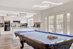 Une table de billard dans une pièce spacieuse, comprenant un coin cuisine, de grandes fenêtres et un ventilateur de plafond.