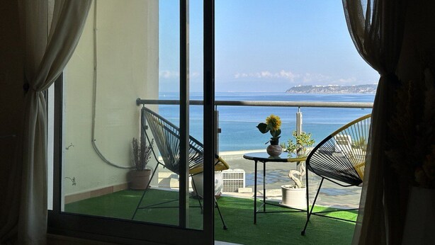 Un balcón con vistas al mar, dos sillas, una mesita con un girasol y una planta en maceta.
