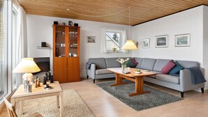 Un soggiorno con soffitto in legno, un divano grigio, un mobiletto in legno, un tavolo con una lampada e una finestra con tende.