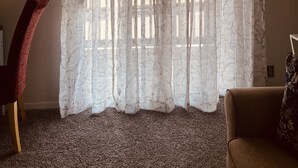 Une pièce avec une grande fenêtre recouverte d’un rideau léger, un fauteuil rouge, un canapé marron et un thermostat fixé au mur.