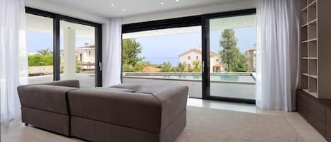Een moderne slaapkamer met een groot bed, schuiframen naar een balkon en uitzicht op zee en gebouwen.