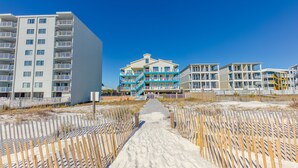 A beachfront hotel with multiple balconies, a central staircase, and a sandy pathway leading to the entrance.