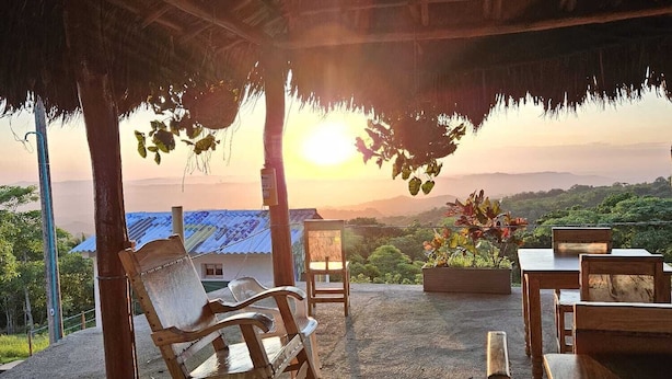 Un área de estar al aire libre con muebles de madera bajo techo de paja, con vista a un paisaje pintoresco al atardecer.