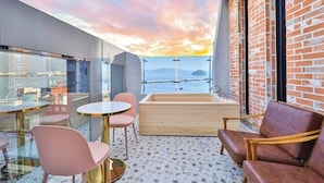 A balcony with a view of the sea, a wooden bathtub, pink chairs, and a round table. - Hotel 701 (Changwon)