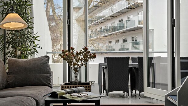 Un salon moderne avec un canapé gris, une petite table sur laquelle reposent un magazine et des lunettes, un vase rempli de fleurs et une grande fenêtre donnant sur un balcon et des immeubles.