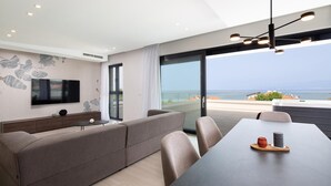 A modern living room with a large TV, a sofa, a dining table with chairs, and a view of the sea.