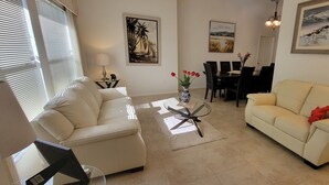 Un salon avec deux canapés, une table basse et une table à manger avec des chaises. Des tableaux ornent les murs et une lampe se trouve sur une table d’appoint.