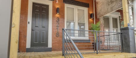 A brick building with a black door, a number 3218, and a wooden porch with a metal railing.