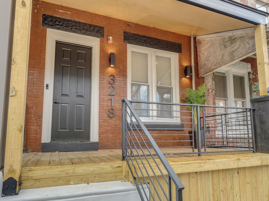 A brick building with a black door, a number 3218, and a wooden porch with a metal railing.