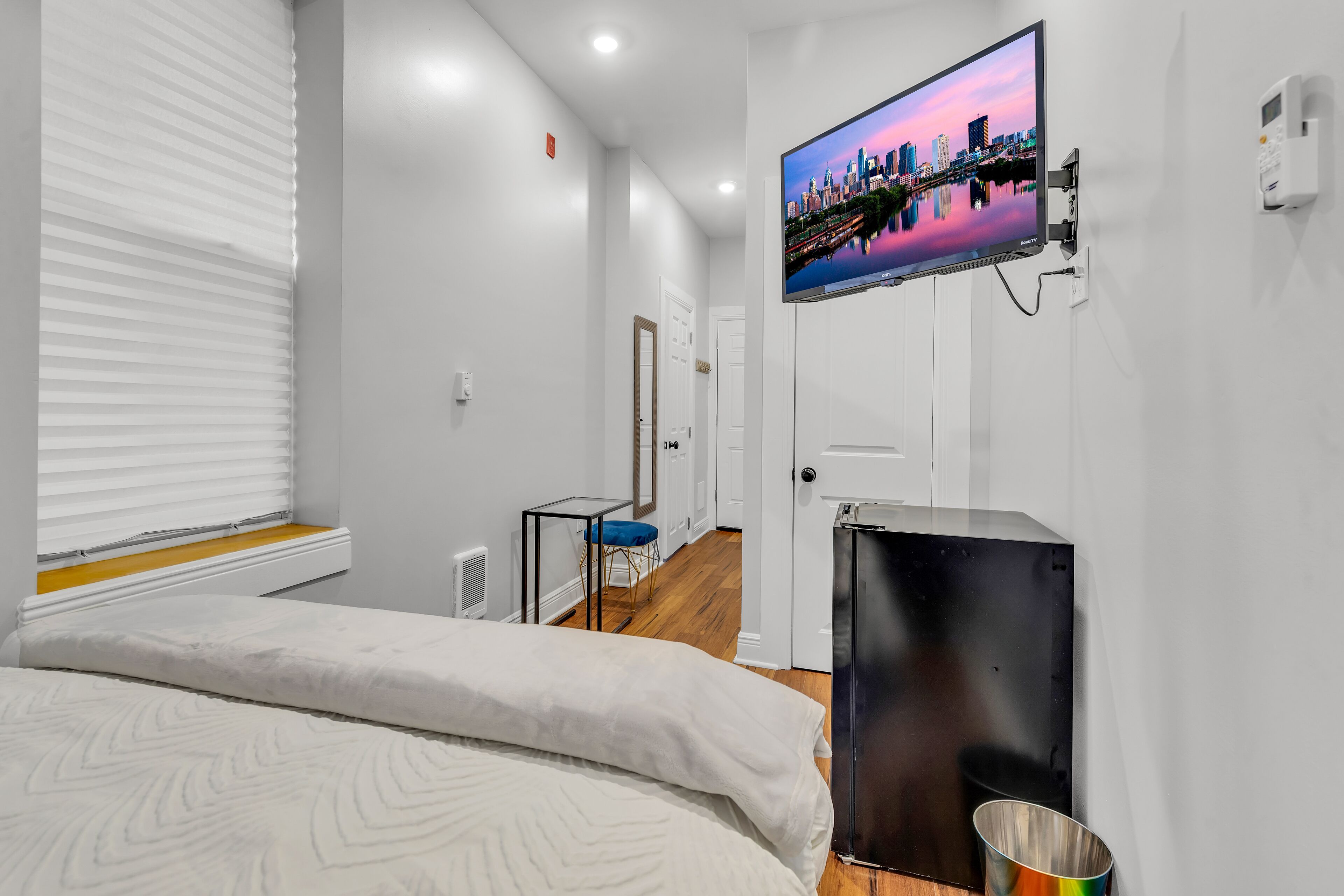 A modern hotel room with a bed, a TV mounted on the wall, a small table, and a door leading to another room.