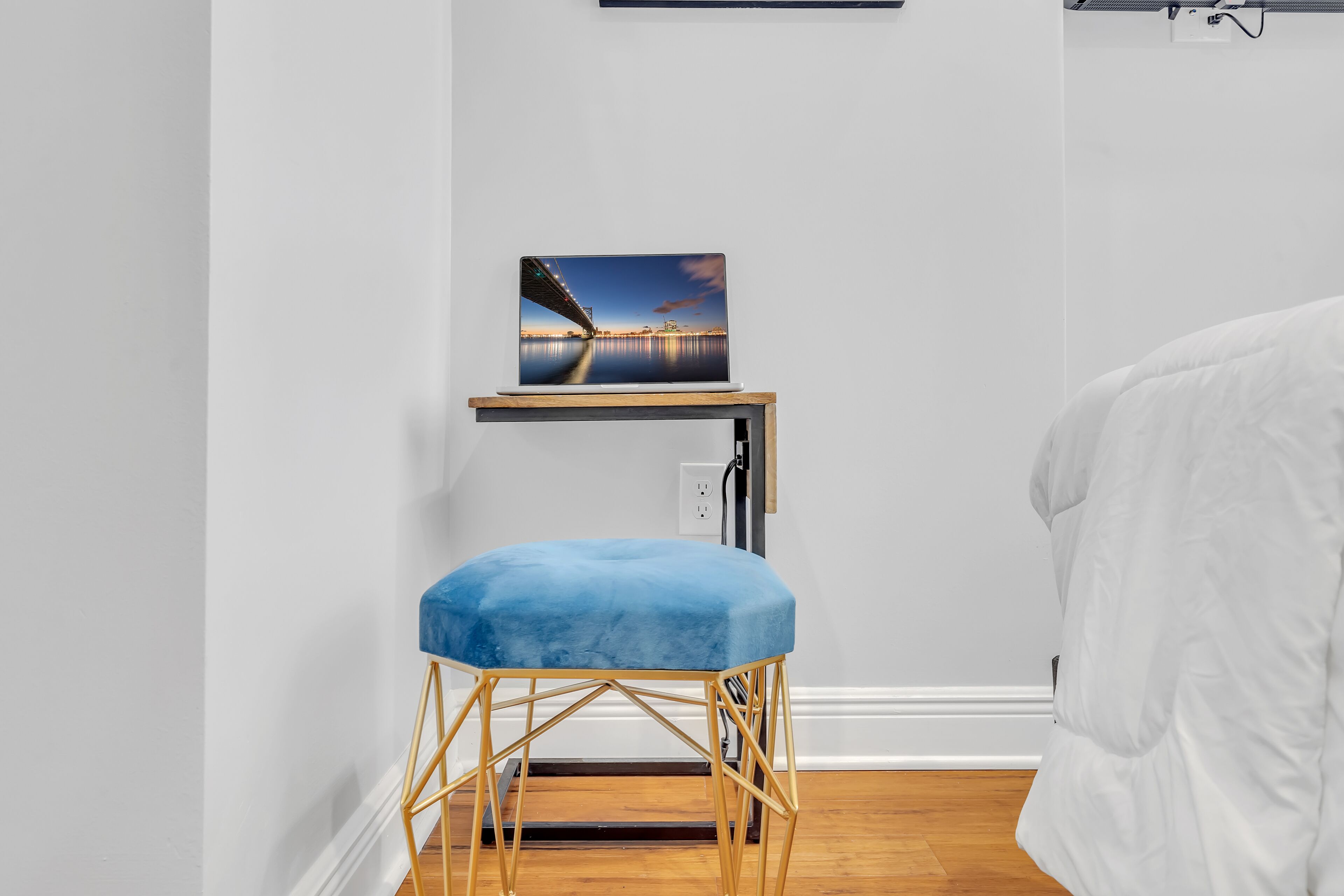 Una habitación con una cama, un taburete azul acolchado, una mesa de madera con un cuadro y una pared blanca.