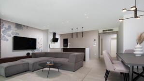 Een moderne woonkamer met een modulaire bank, een salontafel en een aan de muur bevestigde televisie. Er is een eethoek met een tafel en stoelen, en een keuken op de achtergrond.