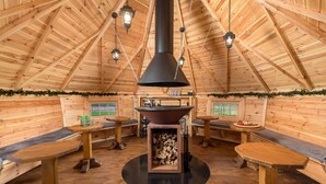 Een gezellig houten blokhut met een houtkachel in het midden, banken, tafels en hangende lantaarns.
