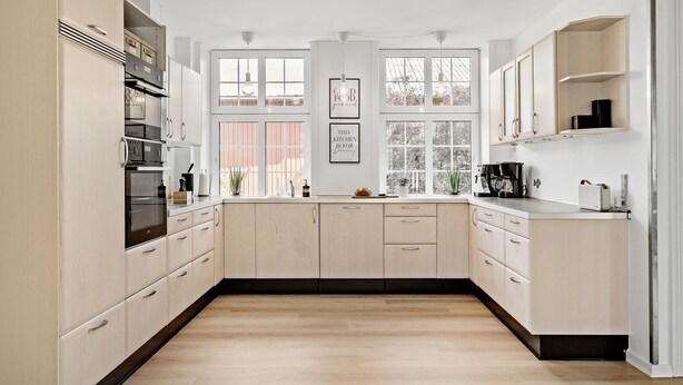 Une cuisine moderne avec des meubles en bois clair, des appareils électroménagers en acier inoxydable et de grandes fenêtres.