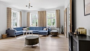 Un salon avec un canapé d’angle bleu, un pouf rond, une petite table d’appoint et une grande fenêtre avec des rideaux.