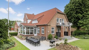 Une maison à deux étages avec une façade en briques, une véranda et une terrasse aménagée avec du mobilier d’extérieur.