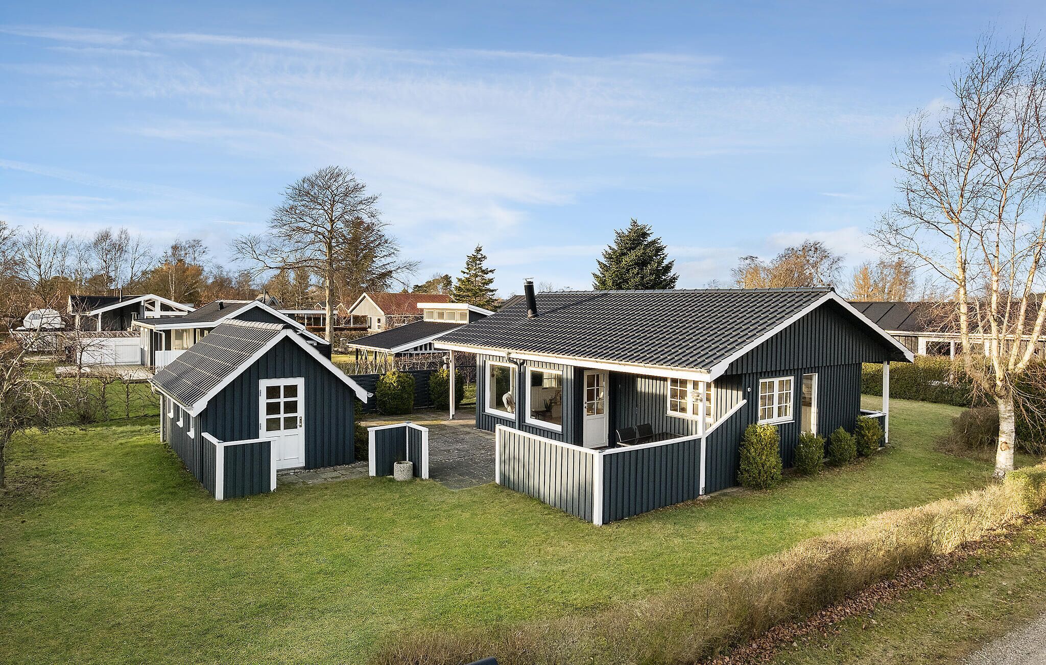 Une rangée de cabanes modernes aux couleurs sombres, rehaussées de bordures blanches, est installée dans une zone herbeuse, avec des arbres et d’autres bâtiments en arrière-plan.