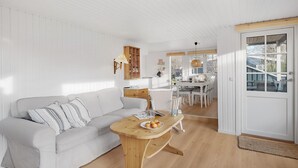 A white-walled room with a wooden floor, a white sofa with striped pillows, a wooden side table with a plate of fruit, and a dining area with white chairs and a table.