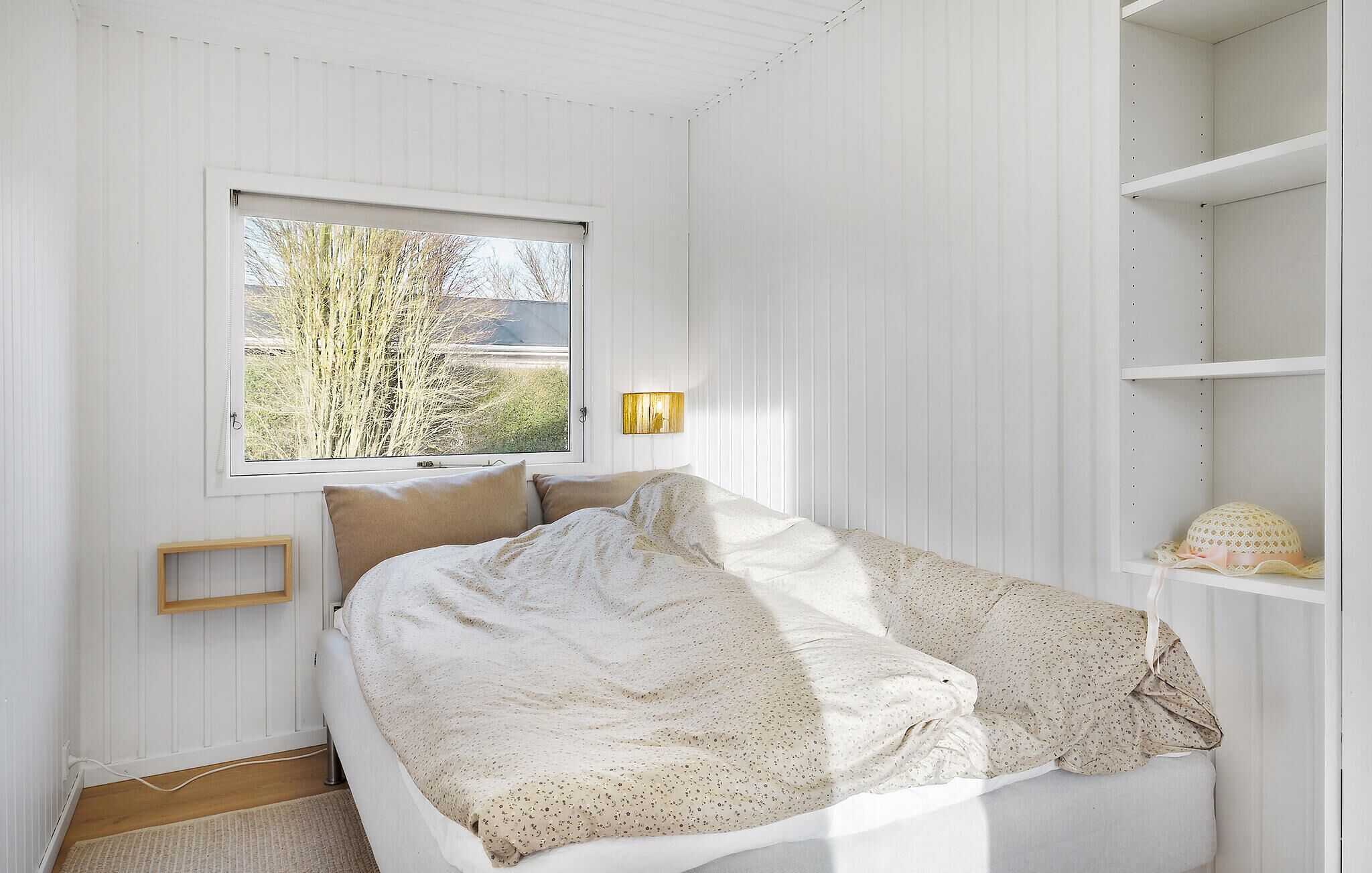 A bedroom with a bed, a window, a wall-mounted lamp, and a shelf with a decorative item.