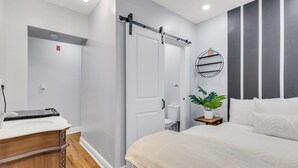 A modern bedroom with a large bed, a wooden dresser, a sliding door, a small bedside table with a plant, and a wall-mounted shelf.
