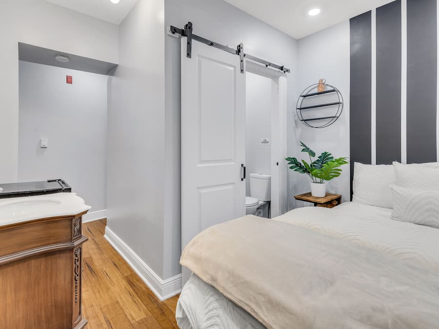 A modern bedroom with a large bed, a wooden dresser, a sliding door, a small bedside table with a plant, and a wall-mounted shelf.