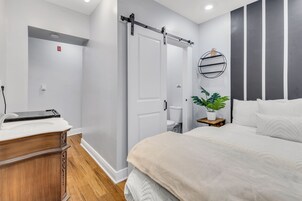 A modern bedroom with a large bed, a wooden dresser, a sliding door, a small bedside table with a plant, and a wall-mounted shelf.