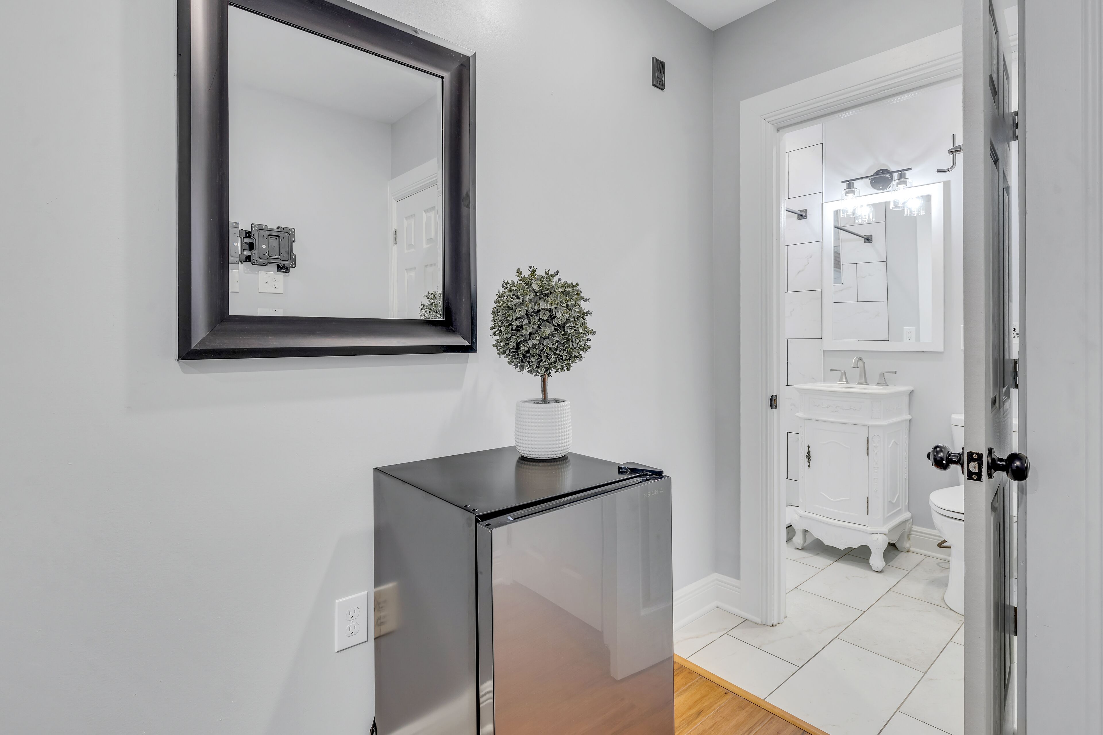 A hallway with a modern black refrigerator, a potted plant, and a white door leading to a bathroom with a white toilet and vanity.