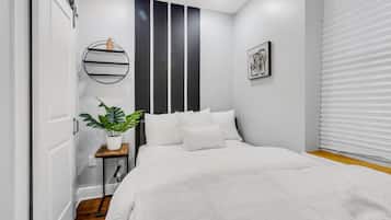 Un dormitorio moderno con una cama grande, una mesita de noche con una planta, un estante en la pared y una ventana con persianas.