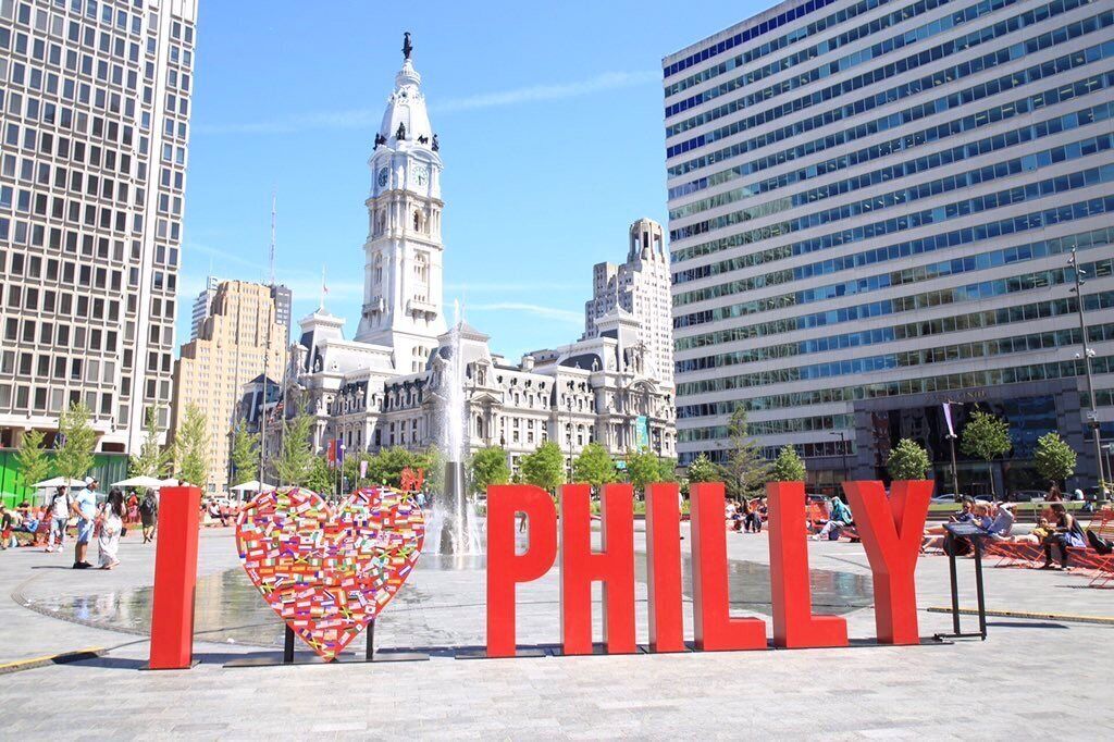 A city square with a prominent 'I ♥ PHILLY' sign, a historic building, and modern skyscrapers in the background.
