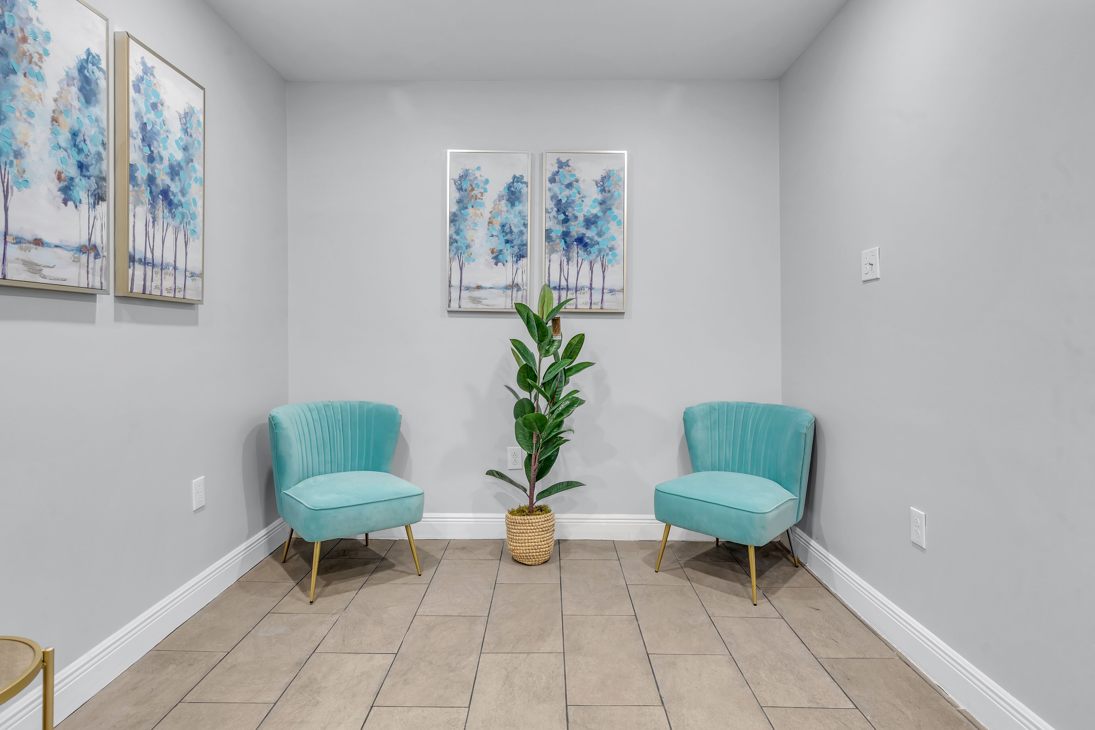 A hallway with two teal armchairs, a potted plant, and two framed paintings on the wall.