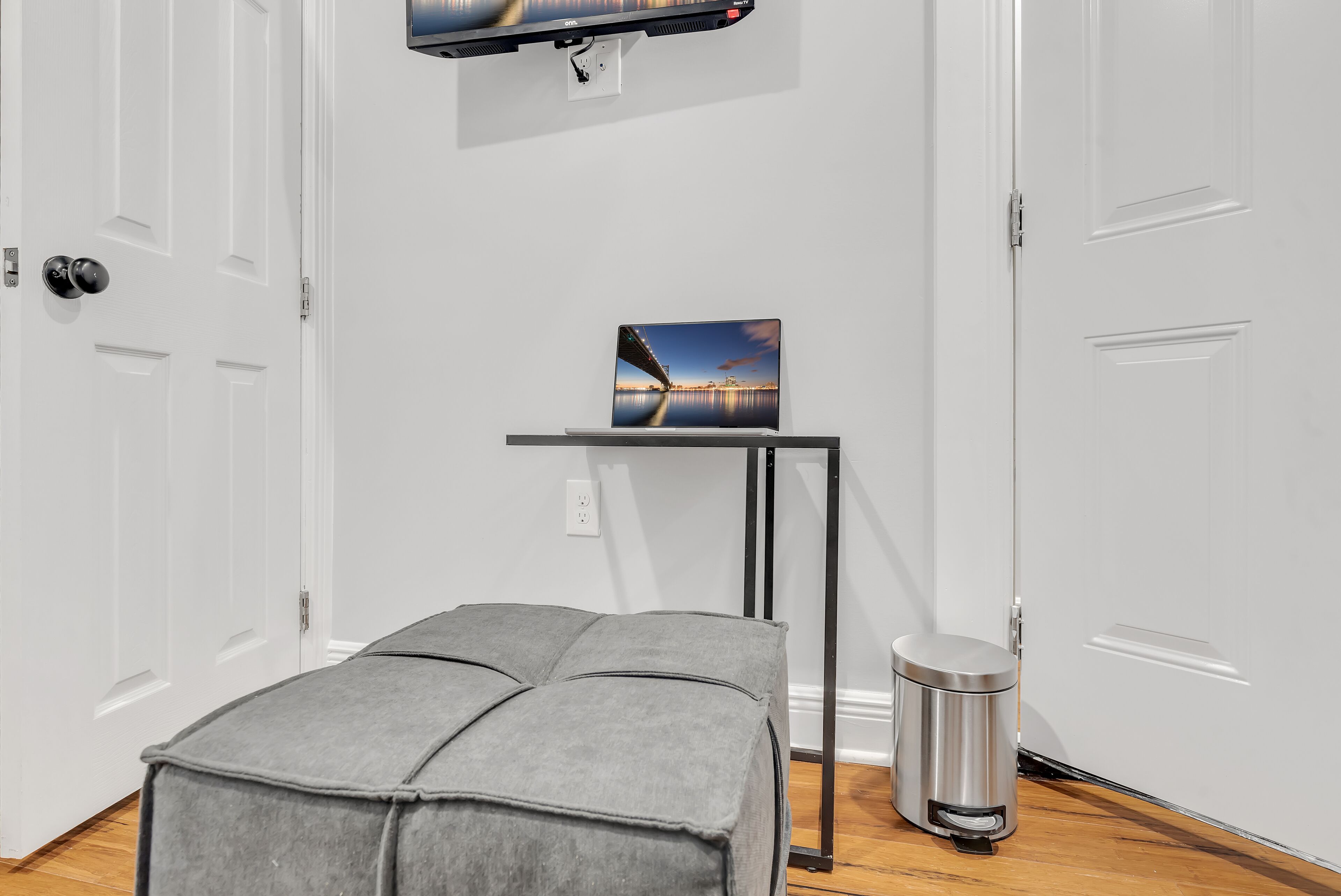 A room with a grey upholstered bench, a small table with a painting, a trash bin, and a television mounted on the wall.