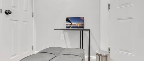 A room with a grey upholstered bench, a small table with a painting, a trash bin, and a television mounted on the wall.