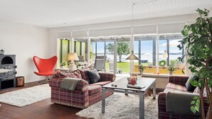 A modern living room with a sofa, armchairs, a coffee table, and a fireplace. Large windows offer a view of the outdoors. - Amazing home in Sunds with lake view (Sunds)