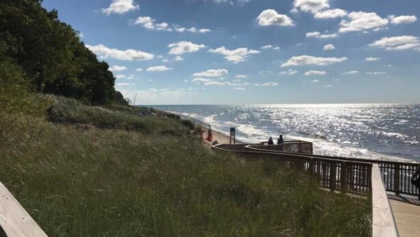 A wooden boardwalk leading to a beach with a clear view of the ocean and a grassy hill on the left.