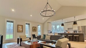 A spacious living room with a large sofa, coffee table, and a chandelier. There is a kitchen area with bar stools and a refrigerator in the background.