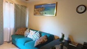 Un salon avec un canapé de couleur turquoise, des coussins à motifs, un tableau représentant un paysage côtier, une horloge et une petite table sur laquelle se trouvent une plante et un livre.