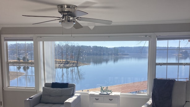 A bedroom with a large bed, two armchairs, a small table, and a ceiling fan. It features a large window with a view of a lake and trees.