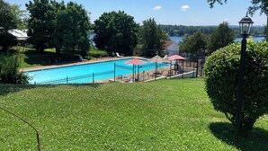 A swimming pool with a fence, surrounded by grass and trees, with a lamp post and a view of a lake.