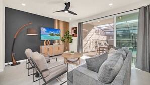 A modern living room with a sofa, chairs, a TV, and a ceiling fan. - Windsor Island Dream | 5BR, 4.5 Bath Cozy Escape w/ Private Pool and Themed Kids' Rooms Near Disney (Davenport)