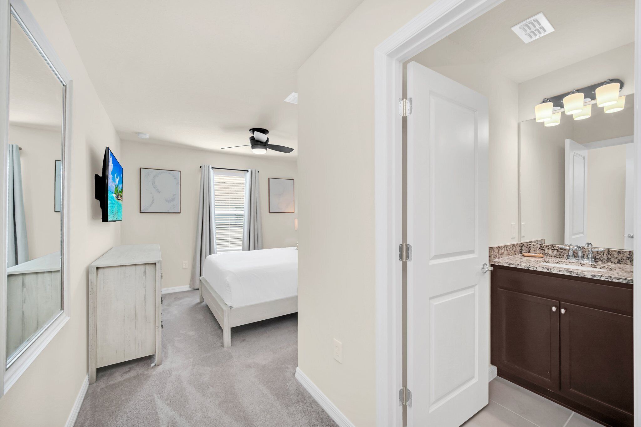 A bedroom with a bed, a ceiling fan, a TV, and a bathroom with a sink and mirror.