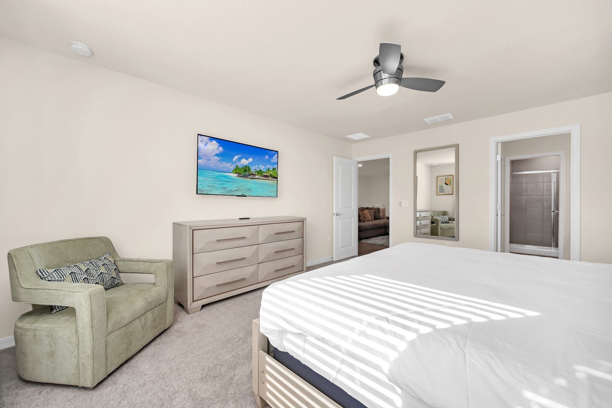 Een slaapkamer met een bed, een groene fauteuil, een dressoir, een plafondventilator en een televisie die een strandscène toont.