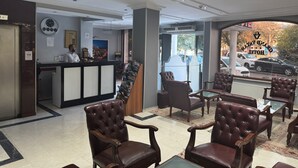 Un hall d’hôtel comprenant un comptoir d’accueil, des fauteuils en cuir, une grande fenêtre donnant sur la rue et une horloge murale.