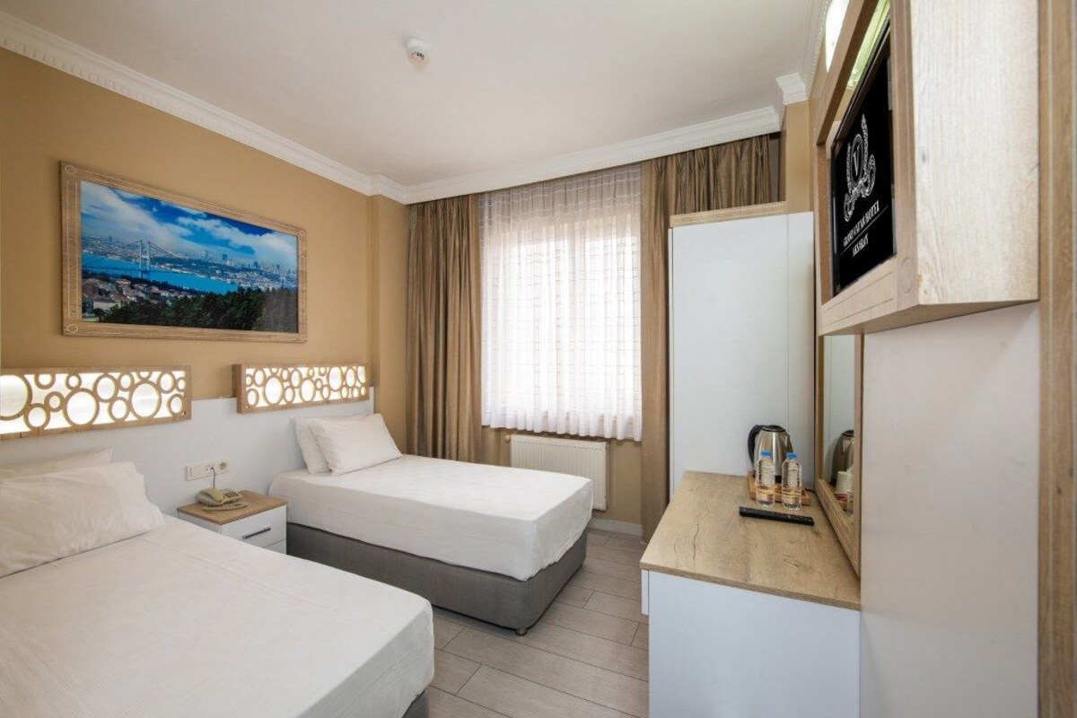 Une chambre d’hôtel avec deux lits, une grande fenêtre avec des rideaux, un tableau représentant un paysage urbain et un téléviseur posé sur un meuble en bois.