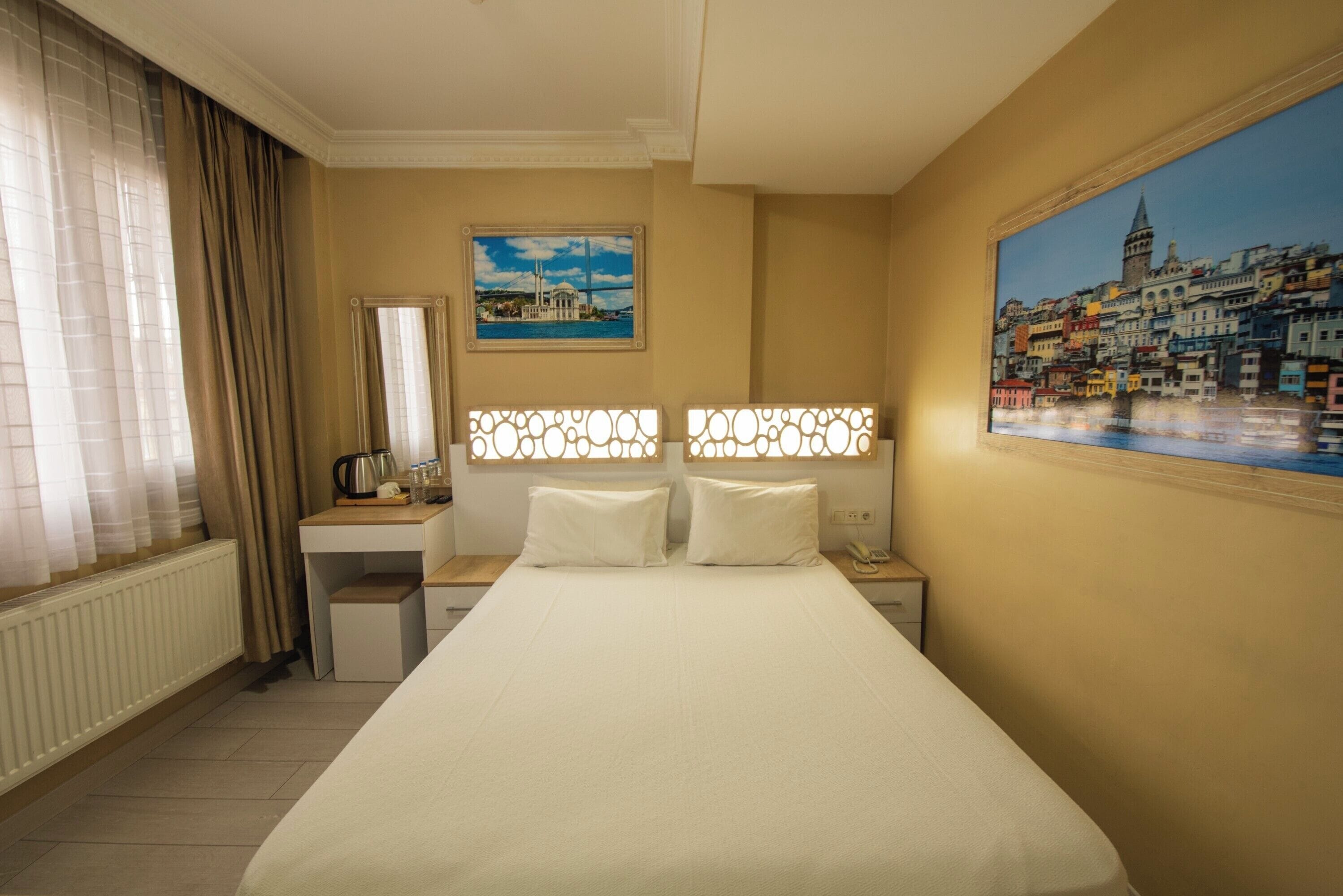 A hotel room with a bed, a desk, a chair, a radiator, a window with curtains, a painting, and a nightstand.