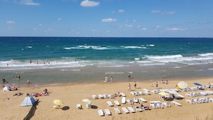 Une plage avec des chaises longues blanches et des parasols, une tente, et un ciel bleu dégagé.