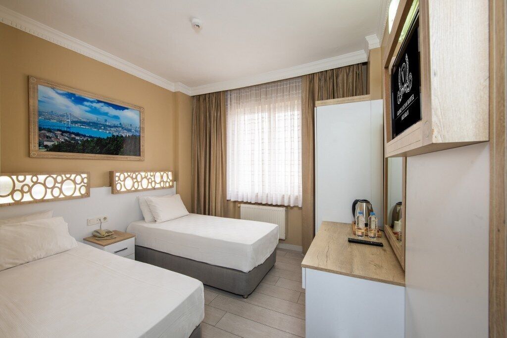 Une chambre d’hôtel avec deux lits, une grande fenêtre avec des rideaux, un tableau représentant un paysage urbain et un téléviseur posé sur un meuble en bois.
