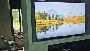 Een moderne kamer met een grote flatscreen-tv aan een groene muur, een houten tafel met stoelen, en uitzicht op een meer door een raam.